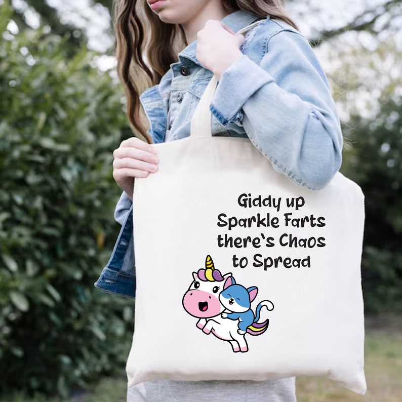 May include: White tote bag with a unicorn and a cat riding on it. The text on the bag reads "Giddy up Sparkle Farts there's Chaos to Spread".