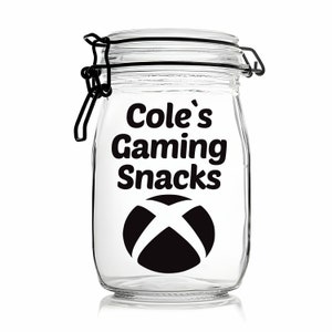 Peut inclure: Un pot en verre transparent avec un couvercle noir et une &eacute;tiquette noire qui dit "Cole's Gaming Snacks" avec un logo Xbox.