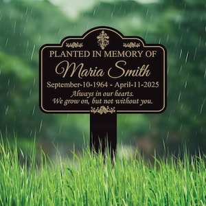 May include: A black and gold memorial garden stake with the text "PLANTED IN MEMORY OF Maria Smith" and dates. The stake also includes the phrases "Always in our hearts" and "We grow on, but not without you."