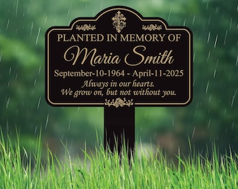 May include: A black and gold memorial garden stake with the text "PLANTED IN MEMORY OF Maria Smith" and dates. The stake also includes the phrases "Always in our hearts" and "We grow on, but not without you."