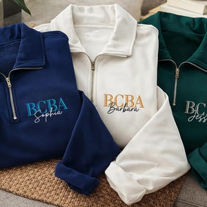 May include: Three quarter-zip sweatshirts in navy blue, white, and emerald green. Each sweatshirt features the embroidered letters "BCBA" with a name below. The navy sweatshirt says "Sophia", the white says "Barbara", and the green says "Jessica".
