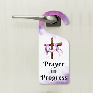 May include: A purple and white door hanger with a watercolor background. A wooden cross with a white cloth draped over it is in the center of the hanger. The text "Prayer in Progress" is printed below the cross.