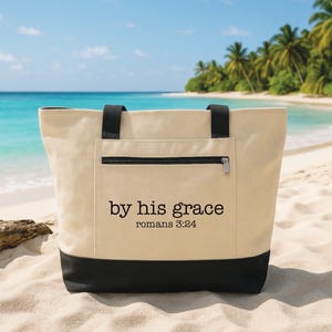 May include: A beige tote bag with black accents, a zippered pocket, and black handles. The bag features the text "by his grace" and "romans 3:24" printed in black. The bag is on a sandy beach with a turquoise ocean and palm trees in the background.