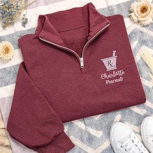 May include: A maroon quarter-zip sweatshirt with a silver zipper. The sweatshirt features a pharmacy-themed logo and the text "Charlotte PharmD" in a light pink color. White sneakers are visible in the bottom right corner.