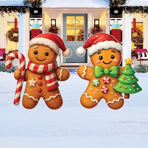 May include: Two gingerbread man yard decorations. One holds a candy cane and wears a red and white striped scarf and Santa hat. The other has a green bow tie and holds a green Christmas tree. Both have smiling faces.