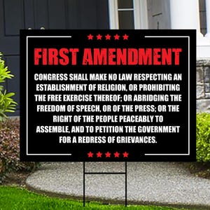 May include: A black and white yard sign with the text "FIRST AMENDMENT" in red. The sign displays the text of the First Amendment to the United States Constitution, with red stars at the top and bottom.