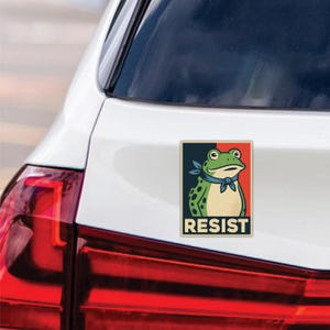 May include: A rectangular sticker featuring a cartoon frog with a blue neckerchief, set against a red, white, and blue background. The word "RESIST" is printed in white block letters at the bottom of the sticker.