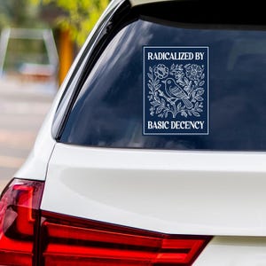 May include: A white sticker on a car window, displaying the text "RADICALIZED BY BASIC DECENCY." The sticker features a detailed white illustration of a bird on a branch, surrounded by flowers and leaves, set against a dark blue background.