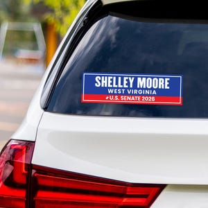 May include: A rectangular sticker on a car window displays the name "SHELLEY MOORE" in white text on a blue background. Below, "WEST VIRGINIA" and "U.S. SENATE 2026" are shown. The sticker has a red stripe at the bottom.