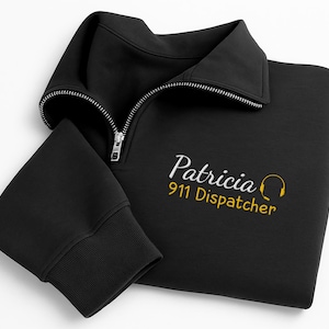 May include: Black quarter-zip sweatshirt with a ribbed cuff and collar. The sweatshirt features the name "Patricia" in cursive script, along with "911 Dispatcher" and a yellow headset graphic.