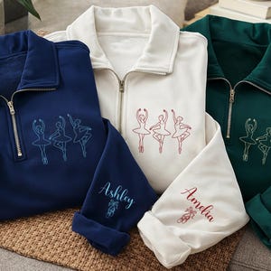 May include: Three quarter-zip sweatshirts in navy blue, cream, and dark green. Each features an embroidered trio of ballet dancers on the chest and a name with ballet slippers on the sleeve. The names are Ashley, Amelia, and Susan in matching thread.