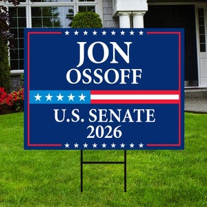May include: A blue campaign sign with the name "JON OSSOFF" in white letters. The sign also includes "U.S. SENATE 2026" and a red, white, and blue graphic. The sign is displayed on a green lawn.
