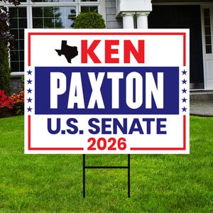 May include: A white and red sign with the text "KEN PAXTON U.S. SENATE 2026" in bold letters. The sign includes a black silhouette of the state of Texas and is displayed on a green lawn with a black metal stake.