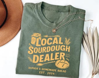 May include: Green t-shirt with a yellow graphic that says "Local Sourdough Dealer" with a jar of sourdough starter, a spoon, and a loaf of bread. The text below the graphic says "Sophia's Homemade Bread Est. 2024".