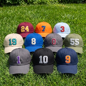 Puede incluir: Una colección de diez gorras de béisbol de varios colores, cada una con un número grande bordado en la parte delantera. Las gorras incluyen granate con 24, naranja con 7, azul claro con 3, beige con 19, azul con 8, gris con 0, oliva con 55, gris oscuro con 1, negro con 10 y azul marino con 9.