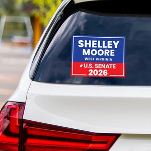 May include: A rectangular sticker on a car window displays the text "SHELLEY MOORE WEST VIRGINIA U.S. SENATE 2026" in white lettering against a blue and red backdrop. The sticker is attached to the rear window of a white car.