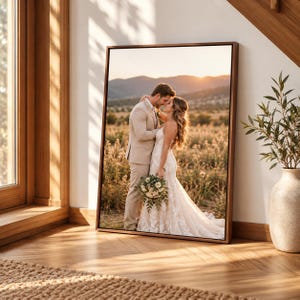 May include: A rectangular framed wall art print featuring a photograph of a couple in wedding attire standing in a field at sunset. The person on the left wears a beige suit, and the person on the right wears a white lace gown holding a bouquet.