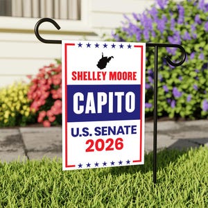 May include: A white and red garden flag with the text "SHELLEY MOORE CAPITO U.S. SENATE 2026" and a silhouette of West Virginia. The flag is displayed on a black metal stand in a grassy garden, with colourful flowers in the background.
