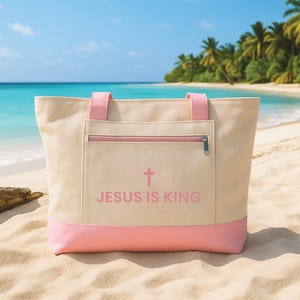 May include: A beige tote bag with pink detailing, including the handles and base, and a front pocket with a zip. The bag displays a pink cross and the text "JESUS IS KING." The background shows a beach with palm trees and the sea.