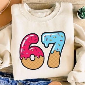 May include: Cream-colored sweatshirt featuring a colorful graphic of the number 67. The numbers are designed to look like donuts, with pink frosting on the 6 and blue frosting on the 7, both with sprinkles and waffle cone bottoms. A fun, playful design.