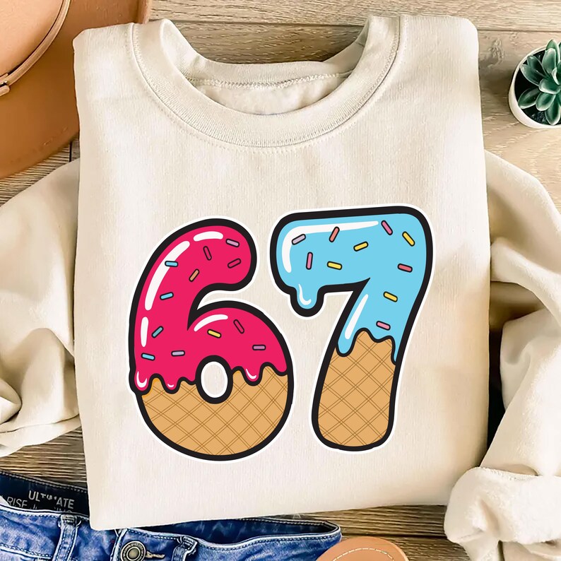 May include: Cream-colored sweatshirt featuring a colorful graphic of the number 67. The numbers are designed to look like donuts, with pink frosting on the 6 and blue frosting on the 7, both with sprinkles and waffle cone bottoms. A fun, playful design.