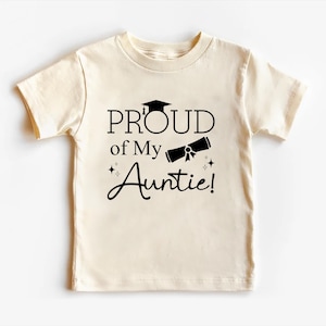 May include: A cream-colored t-shirt with short sleeves features a black graphic that says "Proud of My Auntie!" with a graduation cap and diploma.