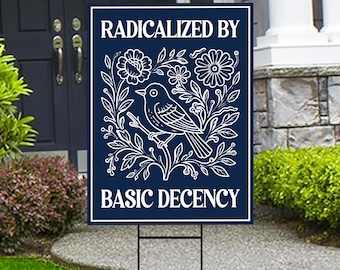 May include: A navy blue yard sign with white text and a floral bird design. The sign reads "RADICALIZED BY BASIC DECENCY". The bird is perched on a branch surrounded by flowers and leaves.