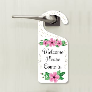 May include: White door hanger with a gold polka dot pattern and pink flowers. The hanger has the text "Welcome Please Come in".