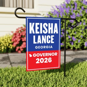 May include: A garden flag with the text "KEISHA LANCE GEORGIA GOVERNOR 2026" in white letters on a blue and red background. The flag is displayed on a black metal stand in a grassy yard, with colorful flowers in the background.