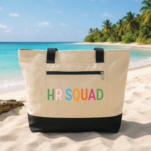 Peut inclure: Un sac fourre-tout en toile beige avec un panneau inférieur et des bretelles noirs. L'avant présente une poche zippée avec le texte HR SQUAD imprimé en lettres multicolores, notamment vert, rose, bleu, orange et rouge sur le tissu beige.