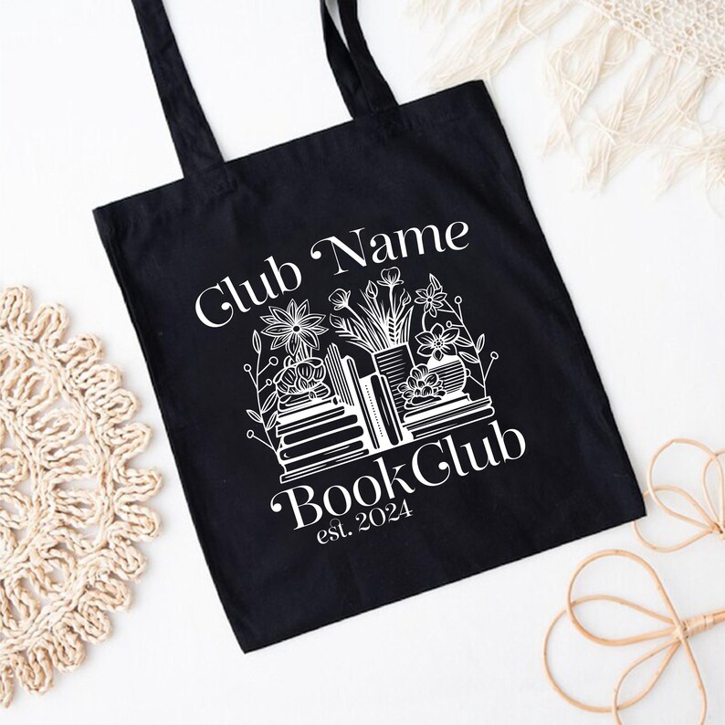 Puede incluir: Bolsa de tela negra con texto blanco que dice "Club Name Book Club est. 2024". El dise&ntilde;o presenta una pila de libros con flores y una maceta.