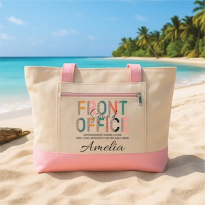 May include: A beige canvas tote bag with pink detailing, displaying the text "FRONT OFFICE Squad" in various colours. The bag has a zipped pocket and pink handles. The name "Amelia" is printed below the text. The bag is on a sandy beach.