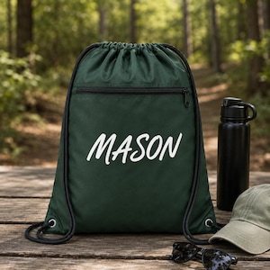 Peut inclure: Un sac à dos à cordon de couleur vert foncé avec une poche zippée horizontale et le prénom MASON imprimé en lettres cursives blanches. Le sac possède des cordons noirs et est placé à côté d'une gourde noire et d'une casquette vert clair.