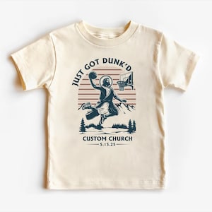 May include: A beige t-shirt with a graphic of a person dunking a basketball. The graphic includes the text "Just Got Dunk'd" and "Custom Church - 5.15.25".