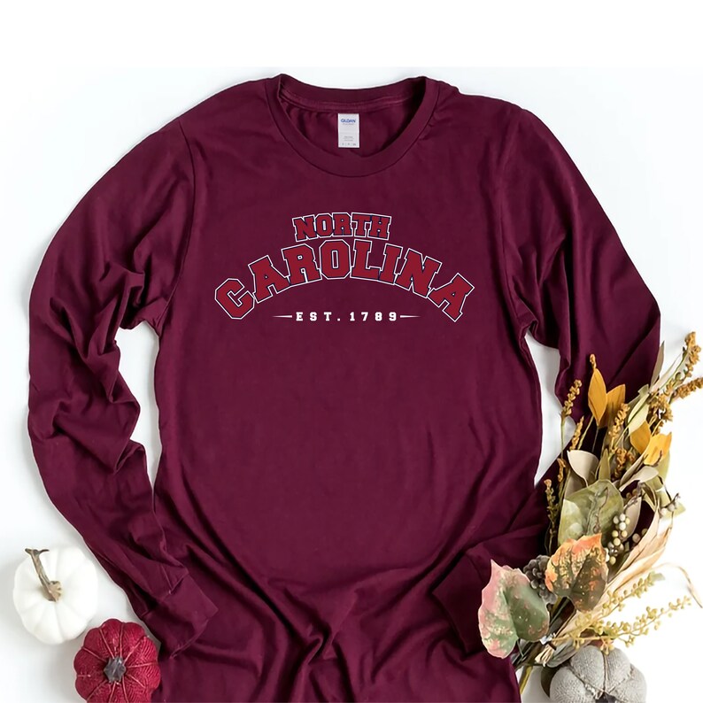K&ouml;nnte beinhalten: Ein lang&auml;rmliges T-Shirt in Bordeaux mit einem wei&szlig;en Grafikdruck, auf dem "NORTH CAROLINA -EST. 1789-" steht.