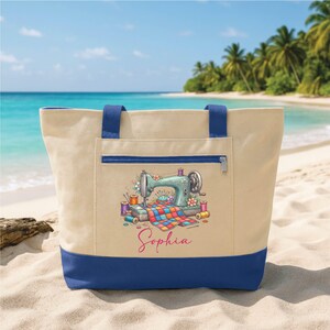 May include: A beige tote bag with a blue bottom and handles, featuring a teal sewing machine graphic with colorful spools of thread and the name "Sophia". The bag has a zippered pocket and is on a sandy beach.