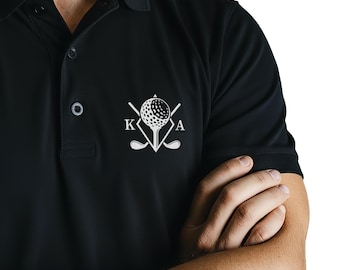 May include: A black polo shirt featuring a white graphic on the chest. The design displays a golf ball on a tee centered between two crossed golf clubs, with the letter K on the left and the letter A on the right, all enclosed in a diamond shape.