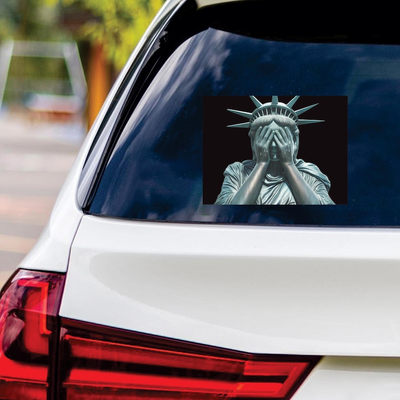 May include: A sticker of the Statue of Liberty with her hands covering her face, set against a black background. The sticker is affixed to the rear window of a white car.