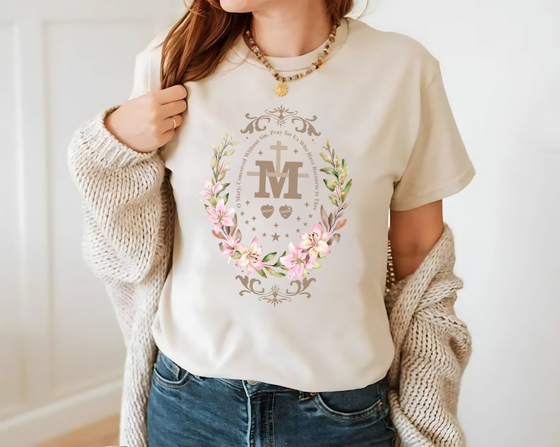 Op de afbeelding: Een beige T-shirt met een bloemenontwerp met een bruine "M" met een kruis en de tekst "O Maria, ontvangen zonder zonde, bid voor ons die onze toevlucht tot u nemen".