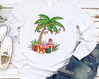 Könnte beinhalten: Weißes Langarmshirt mit einem Weihnachtspalmen-Design. Der Baum ist mit bunten Lichtern und Ornamenten geschmückt. Am Fuß des Baumes liegen Geschenke.