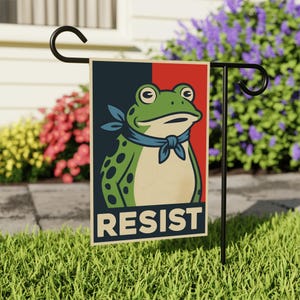 May include: A garden flag featuring a green frog with a blue neckerchief, set against a red and navy blue background. The word "RESIST" is printed in white at the bottom. The flag is displayed on a black metal stand in a grassy yard.