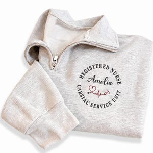 May include: A light grey sweatshirt with a quarter-zip collar. The sweatshirt displays a circular design with the words "REGISTERED NURSE", "Amelia", "CARSIAC SERVICE UNIT", a heart, and a stethoscope.