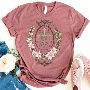 Op de afbeelding: Een mauve t-shirt met een bloemenontwerp met een gouden Maria-kruis en de tekst "Maria, ontvangen zonder zonde, bid voor ons nu en in het uur van onze dood."