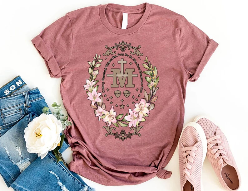 Op de afbeelding: Een mauve t-shirt met een bloemenontwerp met een gouden Maria-kruis en de tekst "Maria, ontvangen zonder zonde, bid voor ons nu en in het uur van onze dood."
