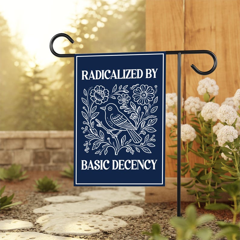 Puede incluir: Una bandera de jard&iacute;n azul marino con el texto "RADICALIZED BY BASIC DECENCY" en blanco. La bandera presenta una ilustraci&oacute;n blanca de un p&aacute;jaro posado en una rama rodeado de flores y hojas. La bandera se muestra en un soporte de metal negro.