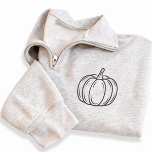 May include: A light gray quarter-zip sweatshirt featuring a black line art illustration of a pumpkin on the front. The garment has a high collar with a silver-toned metal zipper and long sleeves with ribbed cuffs, showcasing a soft fleece interior.