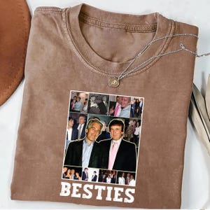May include: A brown t-shirt with a collage of photos and the word "BESTIES" in white. A silver necklace with a pendant is draped on the shirt. The collage features two men in suits.
