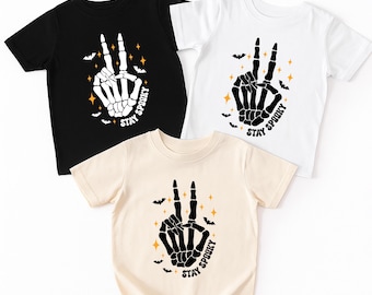 May include: Three short-sleeved t-shirts in black, white, and beige. Each shirt features a skeleton hand in a peace sign, with the words "Stay Spooky" and bat and star graphics. A Halloween-themed design.