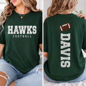 May include: A dark green short-sleeve t-shirt featuring the white distressed text HAWKS FOOTBALL on the front. The back of the shirt displays a brown football graphic above the white distressed vertical text DAVIS in a block font style.
