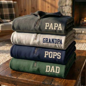 May include: Four quarter-zip pullovers in charcoal, light grey, navy, and forest green are stacked. Each has a white embroidered name: PAPA, GRANDPA, POPS, and DAD. The folded pullovers are on a wooden table. The image is set in a cozy interior.